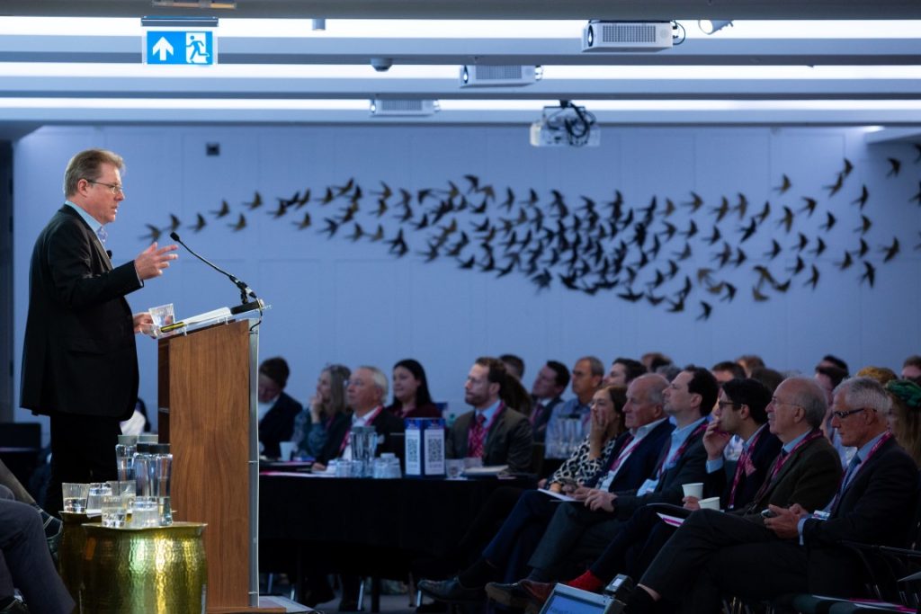 Compass Associates - LaingBuisson Acute Private Healthcare Conference 2022 - speaker stood at podium, side on view of the stage so it views the audieence and the flock of birds displayed on the wall