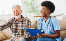 Compass Associates - Residential Care - elderly man sat on a sofa speaking to a healthcare professional with both of them smiling and laughing