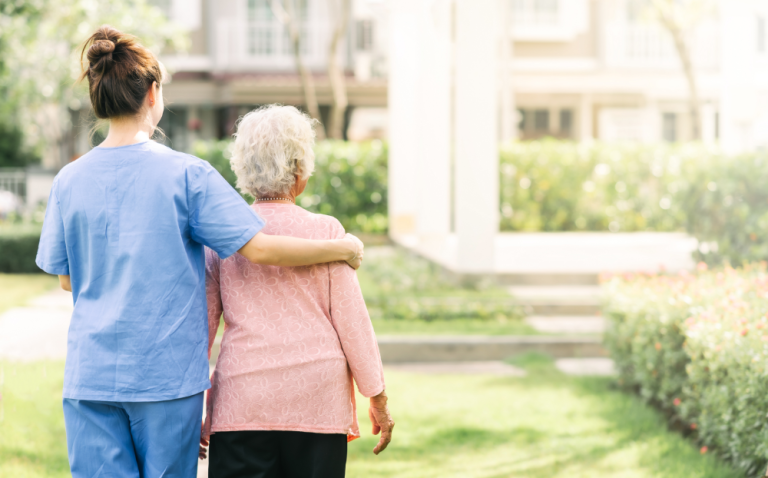 Compass Associates - healthcare / social care professional has arm around elderly lady's shoulder with them both facing away from the camera and looking out over the grass area of a residential provider