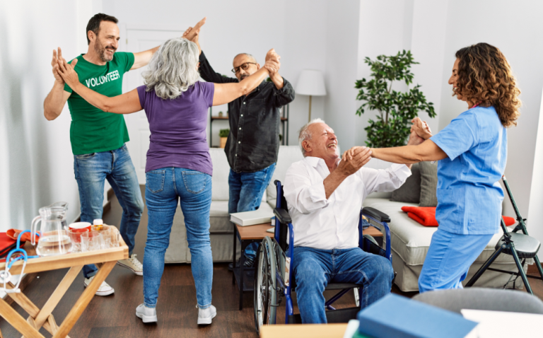 Compass Associates - A group of people happy and dancing in a living room; three people are dancing in a circle while one elderly man is sat in a wheelchair dancing with a female healthcare professional. They are all laughing and smiling.