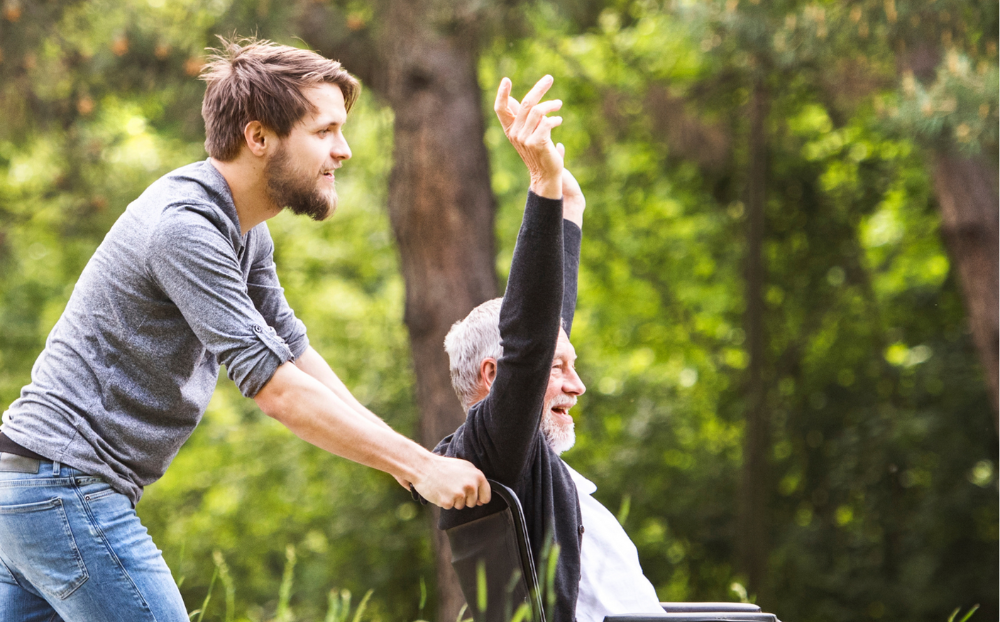 Compass Associates - Young man pushing an elderly man in a wheelchair in an outdoor setting with woodland and greenery behind them. The older man has his arms in the air and they both look happy.