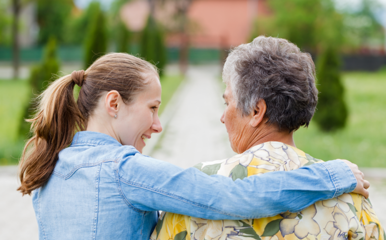 Compass Associates - Two people with their backs to the camera, one with their arm around the other and both looking at each other. They are standing in an outdoor community setting, one is elderly and one is younger.