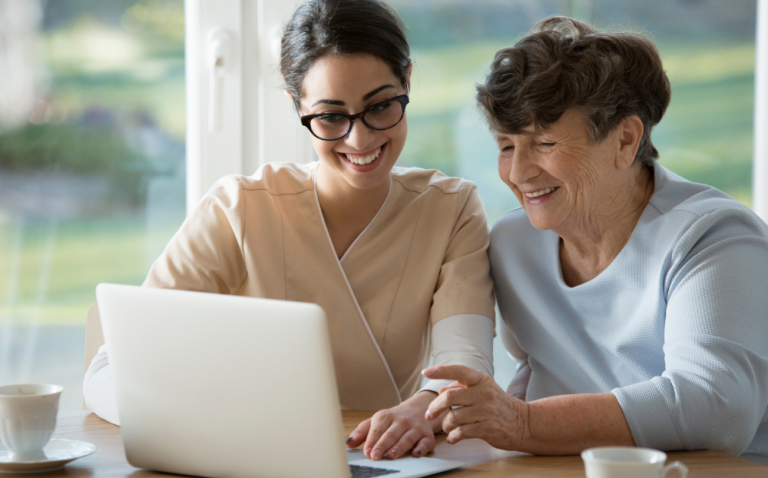 Compass Associates - Two people are sat at a table. One of them is a young lady and one is an elderly lady. They both look happy and smiling, with the young lady showing the other lady a laptop. They also have coffee cups on the table.