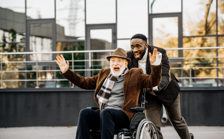 Compass Associates - An elderly man being pushed in a wheelchair by a younger man with both of them looking very happy and enjoying themselves.
