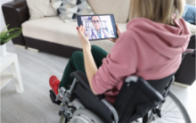 Compass Associates - Healthcare Primary Care - A patient sat in a wheelchair next to a sofa in a living room, holding an iPad that is showing the patient on a video call with a GP