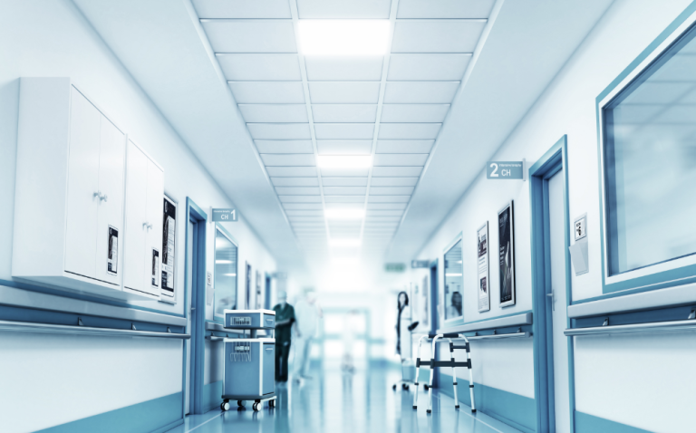 Hospital hallway that has doors to medical rooms, equipment in the hallway and the silhouette of medical professionals in the background