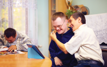 Compass Associates - Specialist Care - Adult Social Care - Three people with learning disabilities sat at a table. One of them is drawing and the other two are laughing and embracing whilse watching a tablet