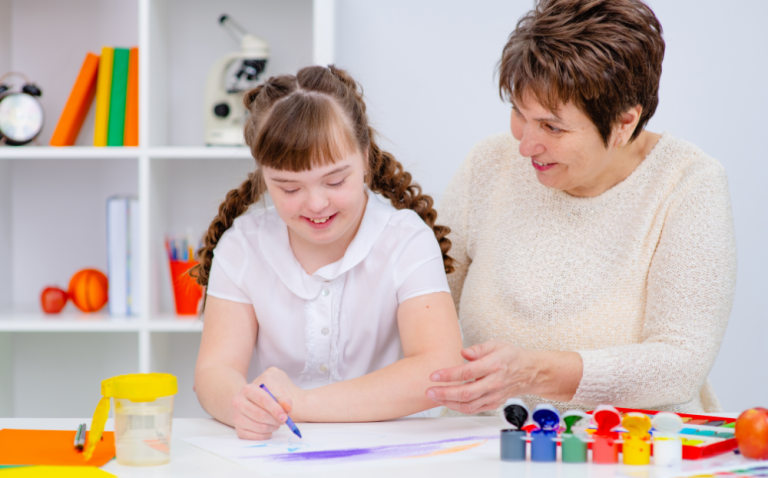 Compass Associates - A teacher / learning assistant supporting a girl with a learning disability with drawing and painting. They are sat at a desk in a classroom setting.