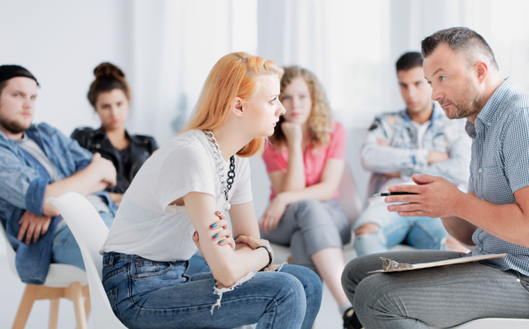 Compass Associates - A young persons support group with four people in the background listening to one person speak with a mental health professional who is also holding a clipboard and pen