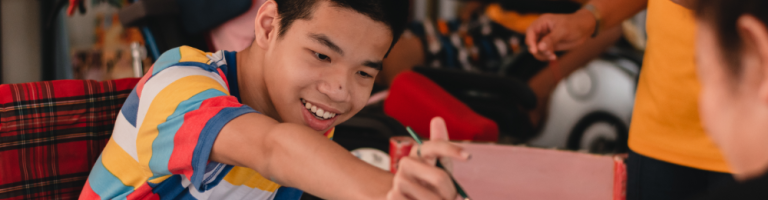 Compass Associates - Specialist Care - Learning Disabilities - A candid photo of a young person with learning disabilities leaning forward and smiling while painting, surrounded by adults and other children playing