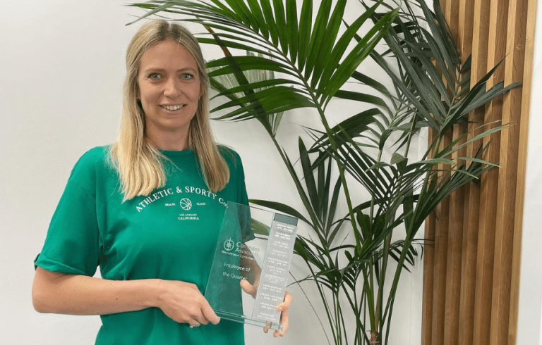 Compass Associates - Natasha Isaac - Employee of the Quarter Q1 - Natasha is posing with her EOQ Award stood by a plant and standing against a white wall. Natasha is wearing a green top and has blonde hair